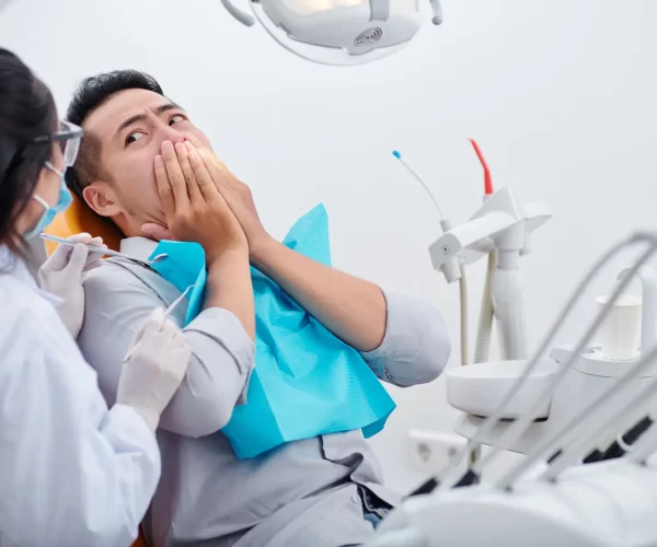 Frightened young Vietnamese man covering mouth and looking at dentist with small mirror and carver