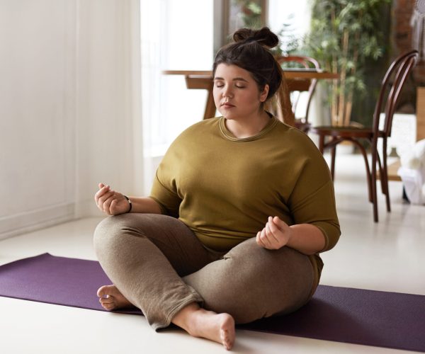 Harmony, balance, zen and peace concept. Indoor shot of plus size barefoot young brunette woman with hair bun sitting on mat keeping legs crossed and eyes shut, meditating after yoga practice