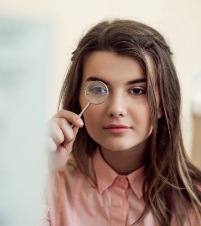 Horizontal portrait of good-looking focused woman on appointment with ophthalmologist holding lense and looking through it while trying to read word chart to check vision. Eyecare and health concept.
