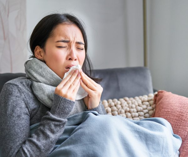 Image of sick korean woman at home, covered in warm clothes and scarf, feeling sick, catching a cold and sitting on sofa sneezing.