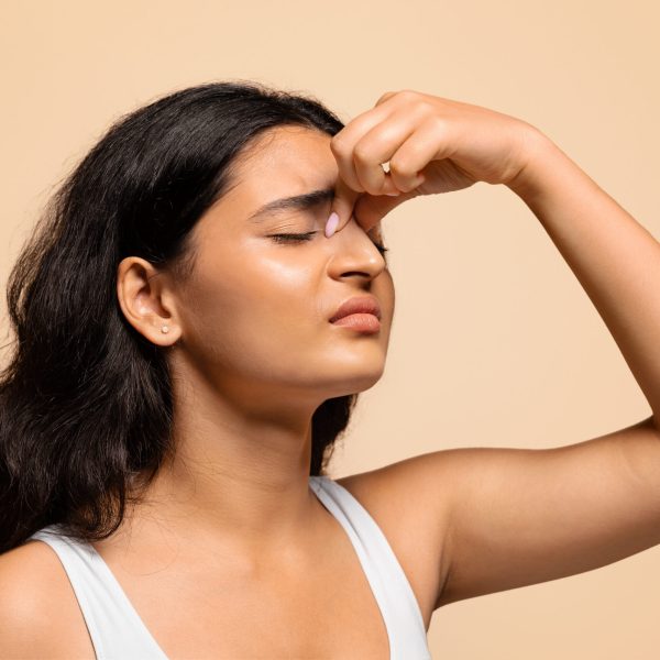Nosebleeds concept. Young indian woman with closed eyes touching nose bridge, upset eastern female suffering from bleeding or dry nose, standing over beige studio background, closeup shot