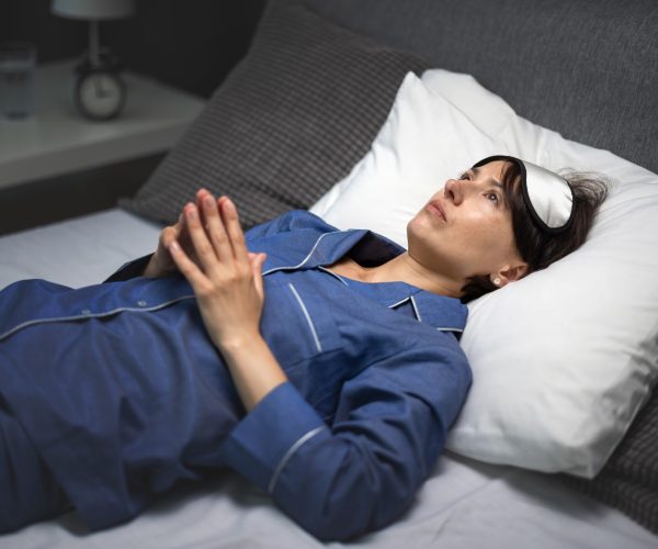 Pensive mature woman in sleep mask and pajamas lying on white soft linen and keeping eyes open during night. Concept of insomnia, problems and sleep disorder.
