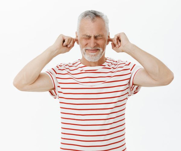 Man closes ears and eyes with intense irritated expression hearing loud disturbing sound waiting till noise disappear standing bothered and annoyed against gray background in striped t-shirt.