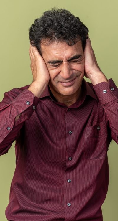 senior man in purple shirt covering eyes with hands with annoyed expression tanding over green background