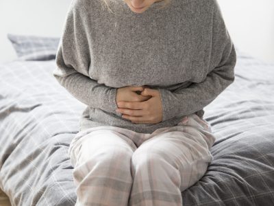 Sick woman in grey homewear sitting on bed, keeping hands on stomach, suffering from intense pain. Illness, stomach ache concept
