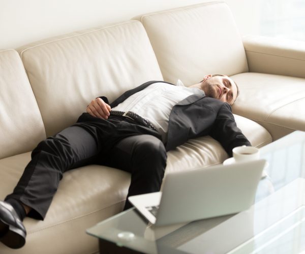 Tired businessman lying relaxed on sofa. Man fall asleep on couch in office when stayed at work till late. Entrepreneur takes short break, recovery sleep after too much hard work at project on laptop