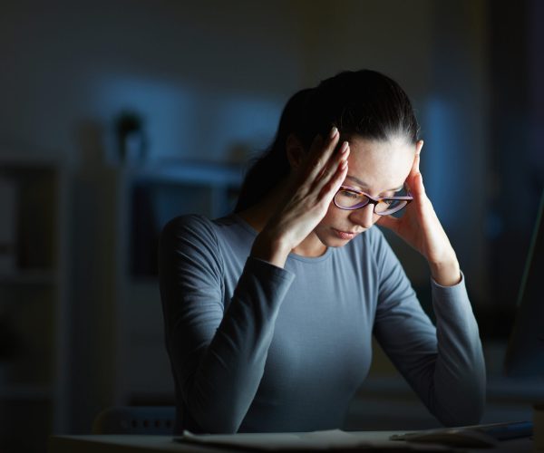 Young businesswoman with headache touching her head while sitting in front of computer monitor by her workplace at night