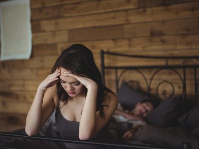 Upset woman sitting on a bed while man sleeping in background at bedroom
