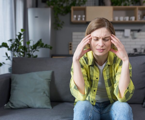 The woman is alone at home, sitting on the couch in the kitchen, has a severe headache