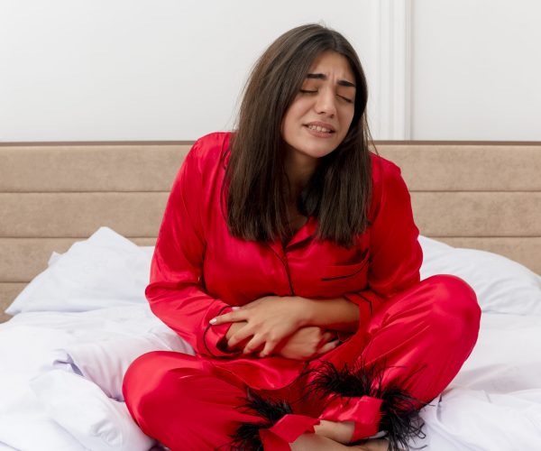 young beautiful woman in red pajamas sitting on bed looking unwell touching her belly suffering from pain in bedroom interior on light background