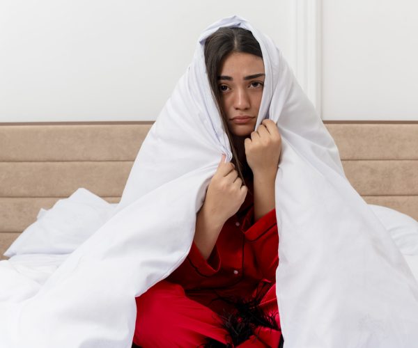 young beautiful woman in red pajamas sitting on bed wrapping in blanket looking at camera with sad expression in bedroom interior on light background